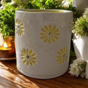 Puede incluir: Un portavelas de cerámica blanca con recortes de margaritas amarillas. El soporte cilíndrico tiene una superficie texturizada y está relleno con un interior amarillo. Las margaritas están espaciadas uniformemente. Una vela encendida es visible en el fondo.