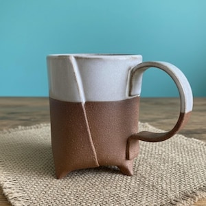 May include: A handcrafted ceramic mug with a unique design. The mug features a two-tone color scheme, with a white upper section and a brown lower section. The handle and base are also brown. The mug is approximately 4 inches tall.