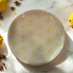 May include: A small, round, ceramic plate with a white background and a pattern of yellow daisies.