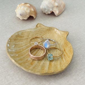 May include: A collection of four rings resting on a textured, tan, shell-shaped ceramic dish. The rings include a wide rose gold band with a brushed finish, a gold band with a small diamond, a silver pear-shaped moonstone ring, and a gold ring.