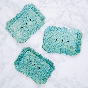 May include: Three turquoise ceramic soap dishes with ornate floral and lace patterns. Each dish has a rectangular shape with a scalloped edge and three drainage holes. The dishes are arranged on a white marble surface.