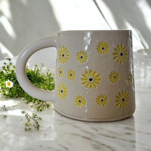 Puede incluir: Taza de cerámica color crema con un asa grande, decorada con diseños de margaritas amarillas. La taza tiene una superficie texturizada y una base redondeada. Las margaritas tienen pétalos amarillos y centros negros. Para bebidas frías o calientes.