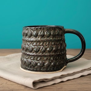 Könnte beinhalten: Eine handgefertigte Keramik-Tasse mit dunkler, strukturierter Außenseite und hellem Boden. Die Tasse hat ein einzigartiges Muster aus erhabenen, abgerundeten Formen und einen dunklen Henkel. Die Tasse steht auf einem neutralfarbenen Tuch.