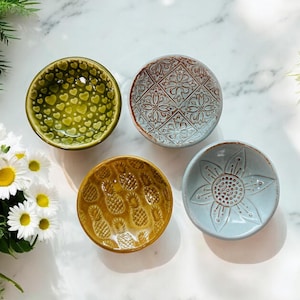 May include: Four small ceramic bowls with different embossed designs. The bowls are green with hearts, blue with a flower, yellow with pineapples, and white with a geometric pattern.