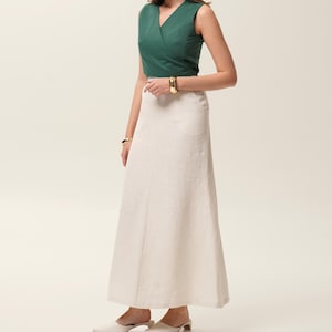 May include: A sleeveless, emerald green wrap top paired with a long, cream-colored skirt. The outfit is accessorized with gold bracelets. The skirt falls to the ankles, and the model is wearing white mules.