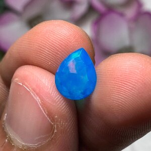 Peut inclure: Une pierre précieuse opale bleue en forme de goutte d'eau et facettée. La gemme est tenue entre les doigts, mettant en valeur sa couleur vibrante et sa taille complexe. La surface de l'opale reflète la lumière, affichant un jeu de couleurs.
