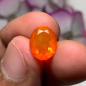 以下が含まれることがあります： オーバルカットの鮮やかなオレンジ色の宝石。指で宝石を持ち、そのファセット加工された表面を見せています。石の色は明るいオレンジ色から、より深い燃えるような色合いまであり、光を反射しています。