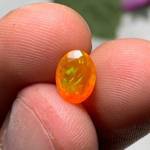 Puede incluir: Gema ovalada y facetada con vibrantes tonos naranja y amarillo. La gema muestra destellos verdes internos, creando un juego de luces. La piedra se sostiene entre los dedos, mostrando su tamaño y brillo. Vista de cerca de las facetas.