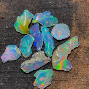 May include: A collection of raw opal gemstones. The opals display a variety of shapes and sizes, with iridescent flashes of color including blue, green, yellow, and orange. The opals are set against a dark, textured surface.