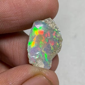 May include: A rough opal gemstone with vibrant flashes of color, including green, red, and yellow. The opal is held between fingers, showcasing its natural, unpolished texture and iridescent play-of-color. The stone has a light, neutral base color.