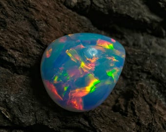 May include: A polished opal gemstone with a rounded shape, displaying a vibrant play of color. The opal features flashes of blue, green, yellow, and red hues. The gemstone is set against a dark, textured background.