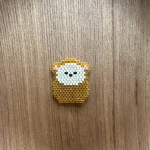Könnte beinhalten: Ein kleiner Affen-Anstecker aus Perlen. Der Anstecker hat einen goldenen Körper, ein weißes Gesicht und rosa Akzente. Das Design ist cartoonartig und detailliert. Der Anstecker ist wahrscheinlich handgefertigt und für Kleidung oder Accessoires geeignet.