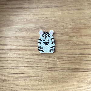 May include: A small, beaded tiger pin with a white body and black stripes. The tiger has small pink ears and a black nose and mouth. The pin is set against a wooden background.