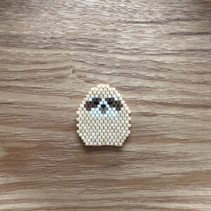 May include: A handmade beaded sloth pin. The sloth is beige with brown and white details, and is made of small, evenly spaced beads. The pin is set against a wooden background.