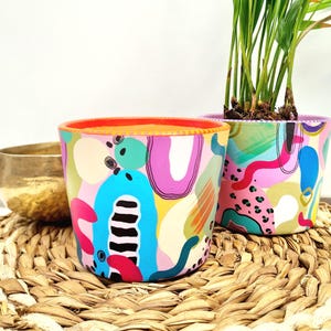 Könnte beinhalten: Zwei handbemalte Keramik-Blumentöpfe mit abstrakten Mustern. Einer hat ein blaues, gelbes und oranges Farbschema, der andere rosa, grüne und violette Farben. Beide Töpfe sind mit grünen Pflanzen gefüllt. Maße in Zentimetern.