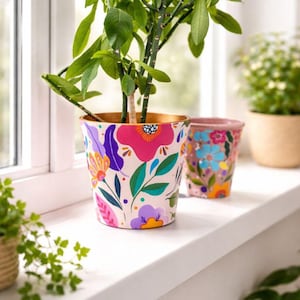 Può includere: Un vaso di fiori in ceramica con un vivace motivo floreale e bordo dorato, contenente una pianta verde. Un vaso più piccolo, simile, con fiori blu è accanto. I vasi sono su un davanzale bianco.