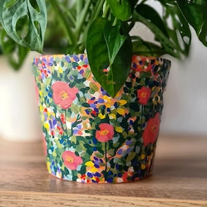 Può includere: Un vaso di fiori dipinto a mano con un colorato motivo floreale con papaveri rossi, accenti blu, gialli e viola. Contiene una pianta verde e poggia su una superficie di legno.