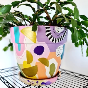 Könnte beinhalten: Ein handbemalter Keramik-Blumentopf mit einem farbenfrohen abstrakten Design. Der Topf weist eine Vielzahl von Formen und Farben auf, darunter Rosa, Gold, Lila und Orange. Der Topf ist mit einer grünen Pflanze gefüllt.