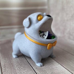 May include: A light grey pug-shaped container with an open mouth, filled with sweets. The pug has yellow eyes, a yellow collar, and a yellow bow tie. It rests on a light-coloured wooden surface.