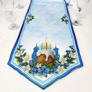 May include: A blue and white table runner featuring a floral design and an illustration of a traditional Easter cake with a candle on top. The cake is surrounded by white flowers and blue flowers.