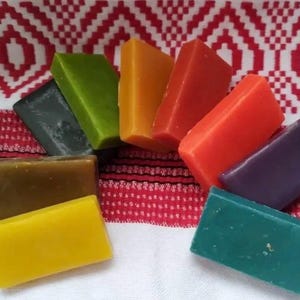 May include: A collection of colorful, rectangular soaps in various shades, including yellow, green, orange, and purple. The soaps are arranged on a white and red patterned cloth, creating a vibrant display. The soaps appear to be handmade.