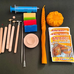 Może przedstawiać: Zestaw materiałów rzemieślniczych do tworzenia zdobionych jajek. Zawiera niebieską strzykawkę, kolorowe bloki wosku, drewniane narzędzia, woskowy element w kształcie słonecznika, wysoką pomarańczową świecę i opakowania z tekstem w języku obcym.