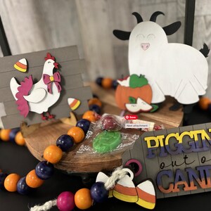 Op de afbeelding: Een houten bord met de tekst "Pigging out on Candy" in kleurrijke letters. Het bord is versierd met een snoepmais en een klein houten kip- en geitenbeeldje. Een reeks oranje en blauwe kralen bevindt zich op de voorgrond.