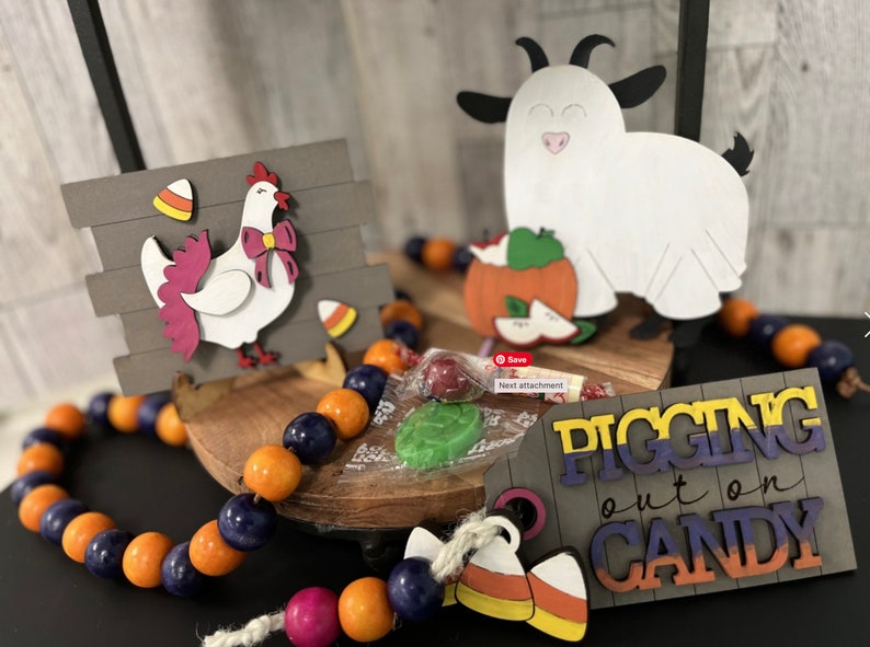 Op de afbeelding: Een houten bord met de tekst "Pigging out on Candy" in kleurrijke letters. Het bord is versierd met een snoepmais en een klein houten kip- en geitenbeeldje. Een reeks oranje en blauwe kralen bevindt zich op de voorgrond.