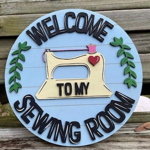 May include: A round, wooden sign with the text "WELCOME TO MY SEWING ROOM." The sign features a light blue background with a cream-colored sewing machine and a red heart. Green leaves and black lettering complete the design.