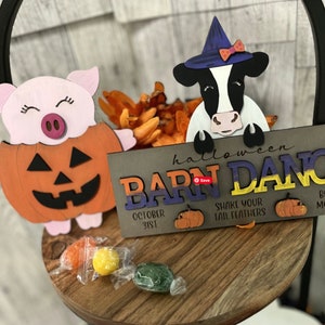 Op de afbeelding: Een houten bord met de tekst "halloween barn dance" in kleurrijke letters. Het bord is versierd met een koe die een heksenhoed draagt en een varken dat een pompoenkostuum draagt. Het bord bevat ook de tekst "October 31st", "Shake your tail feathers" en "Bust a mooove".