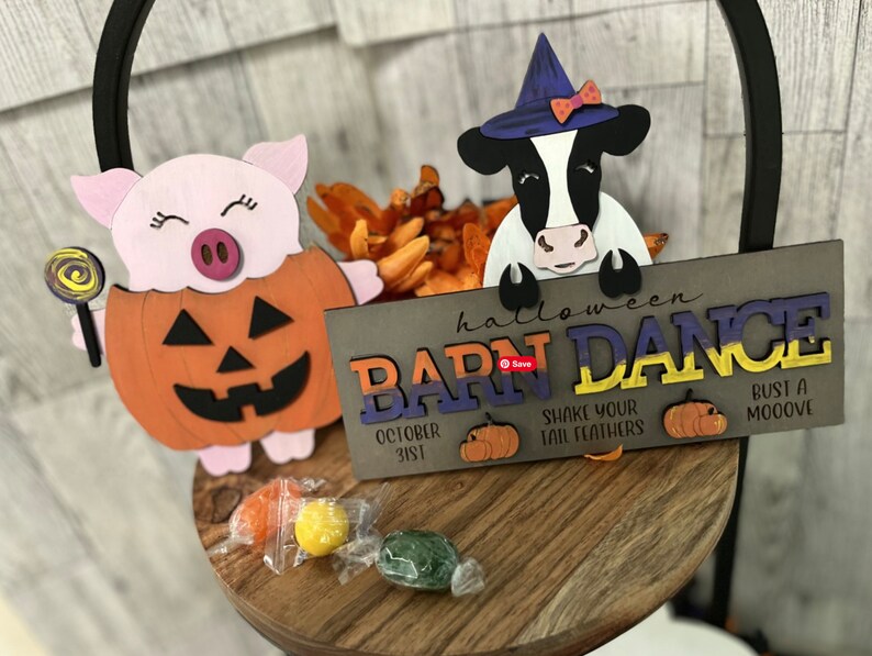 Op de afbeelding: Een houten bord met de tekst "halloween barn dance" in kleurrijke letters. Het bord is versierd met een koe die een heksenhoed draagt en een varken dat een pompoenkostuum draagt. Het bord bevat ook de tekst "October 31st", "Shake your tail feathers" en "Bust a mooove".