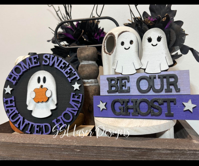 以下が含まれることがあります： ハロウィーンをテーマにした木製装飾。カボチャを持ったお化けと「HOME SWEET HAUNTED HOME」の文字が入った丸い看板。2つのゴーストフィギュアと「BE OUR GHOST」と書かれたブロック。紫と黒のアクセント。