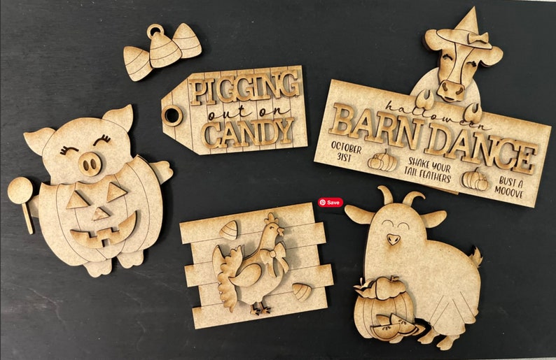 Op de afbeelding: Een set van zes houten uitsnijdingen voor een Halloween-schuurdans thema. De uitsnijdingen omvatten een varken dat een pompoenkostuum draagt, een koe die een heksenhoed draagt, een geit die een pompoen vasthoudt, een kip, een bord met de tekst "Pigging out on Candy", en een bord met de tekst "Halloween Barn Dance".