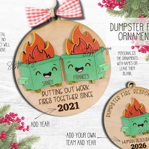 May include: A round wooden Christmas ornament featuring two green dumpster characters with smiling faces and orange flames on top. The text reads: "PUTTING OUT WORK FIRES TOGETHER SINCE 2021" with names "SANDY" and "FRANCES" on the dumpsters. A red and white gingham bow is attached at the top.