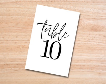 Könnte beinhalten: Eine weiße Tischkarten mit dem Wort "table" in kursiver Schrift und der Zahl "10" in fetter, serifenloser Schrift. Die Karte liegt auf einer hellbraunen Holzoberfläche.