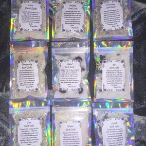 May include: Nine small, clear, iridescent-edged plastic pouches containing bath salts. Each pouch features a white rectangular label with black text identifying the variety: Love, Healing, Protection, Prosperity, Spirit, Air, Fire, Water, and Earth.