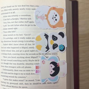 May include: Three colourful, cartoon-style bookmarks on a book page. The bookmarks feature a Shiba Inu, a panda with a flower, and a girl with a butterfly. The bookmarks are designed to mark pages in a book.