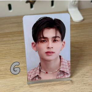 May include: A clear acrylic photo stand displaying a portrait of a person with dark hair, wearing a pink and white plaid shirt and a beaded necklace. The stand is on a wooden surface. A gray letter "G" is visible.