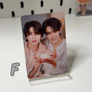 May include: A rectangular photo card featuring two men holding a heart-shaped object. One man wears a pink shirt, the other a white shirt. The card is displayed in a clear acrylic stand.