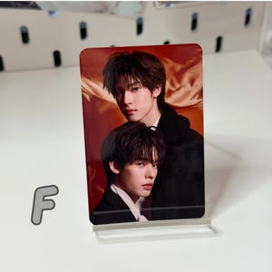 May include: A rectangular photo card featuring two young men in formal attire. The card has a glossy finish and is displayed in a clear acrylic stand. The background is a warm, reddish-orange color.