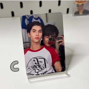 May include: A clear acrylic photo stand holding a printed image of two people. One person is wearing a red and white baseball style shirt with a graphic. The other person is taking a selfie with a phone.