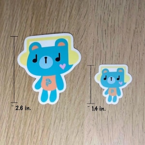 May include: Two stickers featuring a cartoon bear wearing headphones. The larger sticker is 2.6 inches tall, and the smaller one is 1.4 inches tall. The bear is blue with a yellow headset, a pink heart, and an orange letter "P" on its chest.