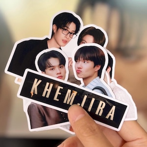 Op de afbeelding: Een verzameling stickers met afbeeldingen van jonge mannen. De stickers zijn in verschillende poses en uitdrukkingen. Eén sticker heeft de tekst "KHEM JIRA" in witte letters op een zwarte achtergrond. De stickers worden door een hand vastgehouden.