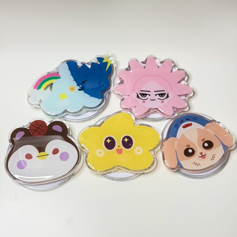 May include: A collection of five clear acrylic pins with cartoon character designs. The pins feature a cloud with a rainbow, a pink sun, a brown penguin with a basketball, a yellow star, and a dog with a shark fin.