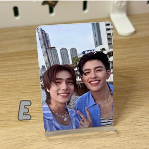 May include: A clear acrylic photo stand displaying a color photograph of two smiling people. The photo shows two men posing for a selfie with a cityscape in the background. The letter "E" is visible on the left side.