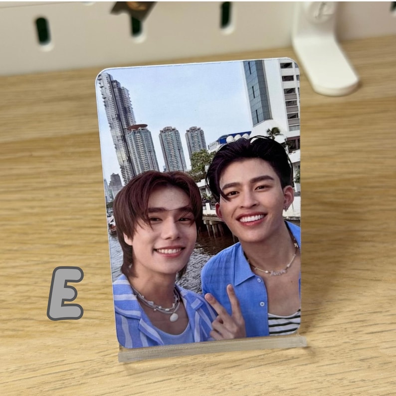 May include: A clear acrylic photo stand displaying a color photograph of two smiling people. The photo shows two men posing for a selfie with a cityscape in the background. The letter "E" is visible on the left side.