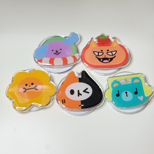 May include: A collection of five colorful acrylic phone grips with cartoon animal designs. The grips feature a dog in a floatie, a cat with a tomato, a sunflower, a cat with a split face, and a bear with headphones.