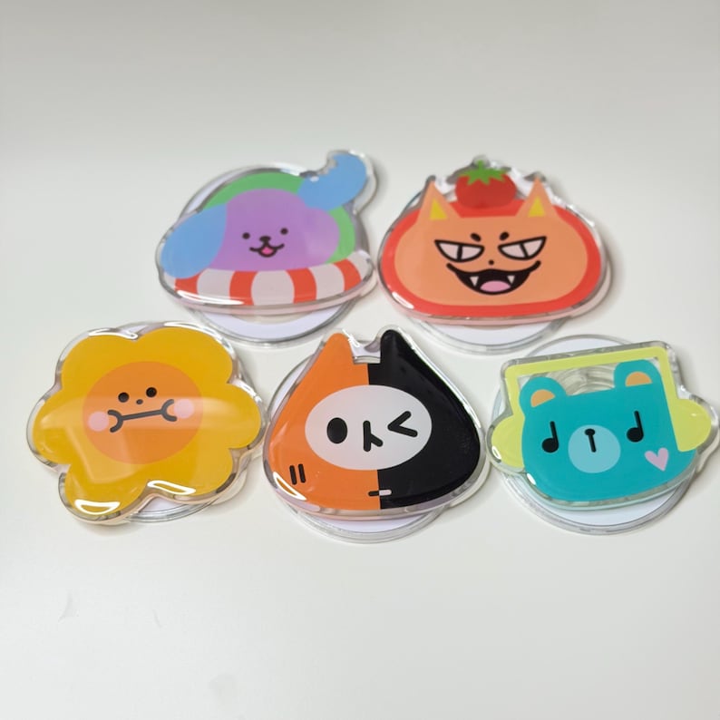 May include: A collection of five colorful acrylic phone grips with cartoon animal designs. The grips feature a dog in a floatie, a cat with a tomato, a sunflower, a cat with a split face, and a bear with headphones.