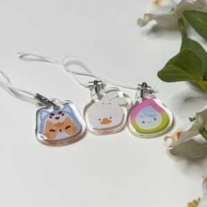 Op de afbeelding: Drie heldere acryl bedels met cartoonontwerpen. Eén heeft een kat met een blauwe hoed, een andere een witte eend en de laatste een roze en geel karakter. Elke bedel heeft een zilveren sluiting en een wit koord.