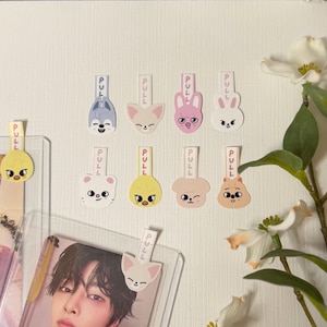 Pode incluir: Uma coleção de onze abas de puxar coloridas com rostos de animais de desenho animado. Cada aba tem um rosto de animal diferente, incluindo um lobo, uma raposa, um coelho e um pintinho. A palavra "PULL" é impressa em cada aba. Algumas abas estão presas a capas de plástico transparente.